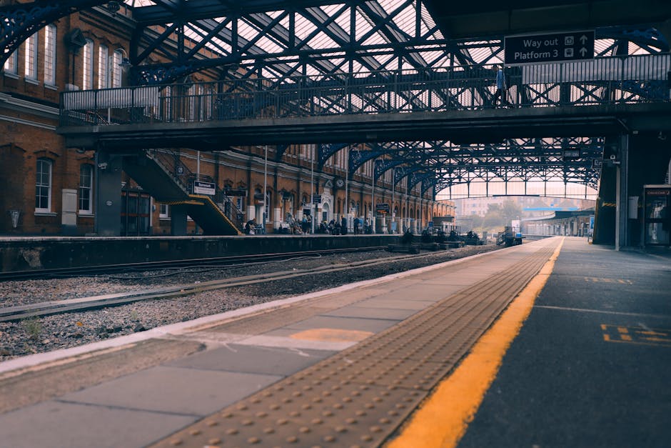 View of West Brompton station platform showing a paved tactile walking surface with yellow safety lines along the edge, under a metal and glass canopy structure supported by blue-painted steel beams. 